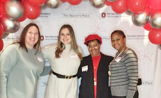Four women in business casual dress stand under a scarlet and gray balloon arch and pose for the picture.